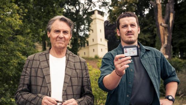 Zwei Männer stehen vor einem Turm, wobei einer von ihnen einen Polizeiausweis in die Kamera hält.