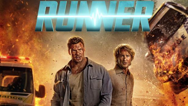 Alan Ritchson und Owen Wilson stehen vor brennenden und explodierenden  Autos, während sie einen Kühlbehälter für Organtransporte tragen.