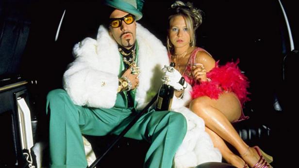 Ein Mann in grünem Anzug mit weißem Pelzmantel und eine Frau in einem pinken Kleid sitzen mit einer Champagnerflasche in einer Limousine.