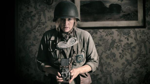 Kate Winslet trägt als Kriegskorrespondentin Lee Miller einen Helm und hält eine Kamera vor dem Hintergrund einer gemusterten Tapete.