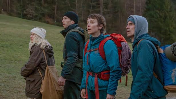 Vier Wanderer mit Rucksäcken stehen auf einer Wiese und blicken mit besorgten Mienen in die Ferne.
