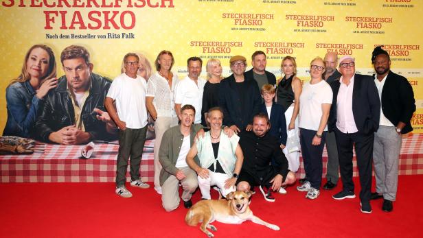 Gruppenfoto auf dem roten Teppich vor dem Filmplakat „Steckerlfisch Fiasko“, mit mehreren Prominenten und einem liegenden Hund.