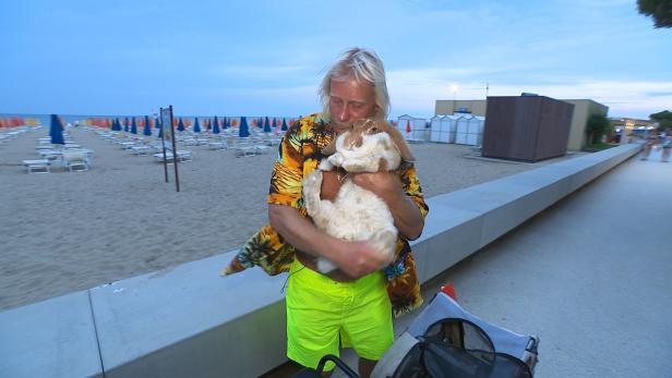 Ein Mann mit buntem Hemd und neongelber Badehose hält ein großes Kaninchen auf dem Arm an einem Strand mit Liegestühlen.