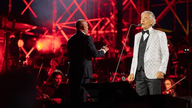 Andrea Bocelli singt vor einem Orchester, während ein Dirigent den Auftritt vor einer rot beleuchteten Bühne leitet.