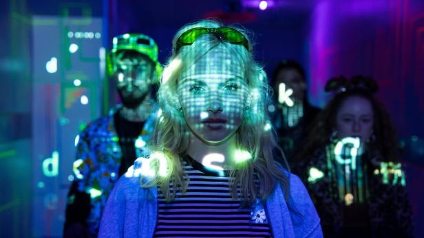 Eine junge Frau mit Brille auf dem Kopf steht im Vordergrund, während auf ihr Gesicht und ihre Begleiter grüne Lichtprojektionen fallen.