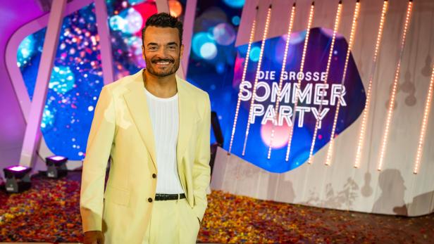 Giovanni Zarrella lächelt in einem hellgelben Anzug vor dem Bühnenhintergrund der Sendung „Die große Sommerparty“.