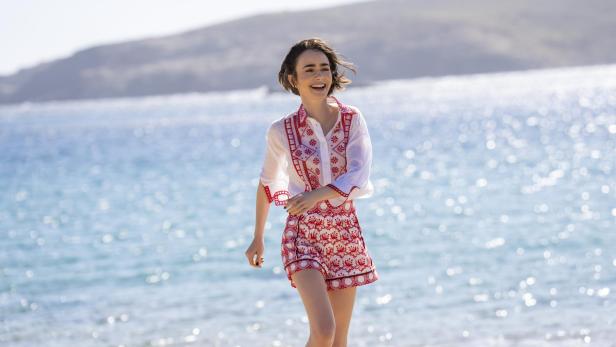 Lily Collins lacht fröhlich, während sie in einem weißen Outfit mit rotem Muster am sonnigen Strand entlangläuft.