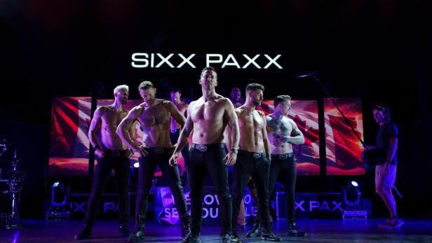 Die Tanzgruppe Sixx Paxx präsentiert sich mit oberkörperfreien Männern auf einer beleuchteten Bühne vor einer kanadischen Flagge.