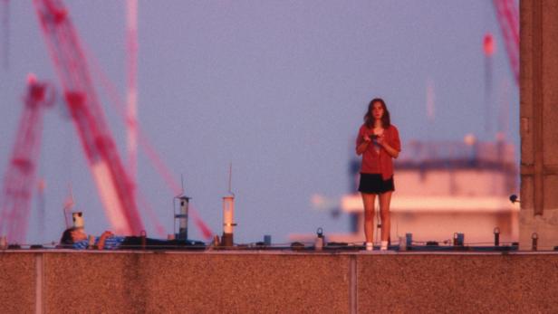 Eine junge Frau steht auf einem Dach, während eine Person daneben liegt, vor dem Hintergrund von Kränen im Abendlicht.