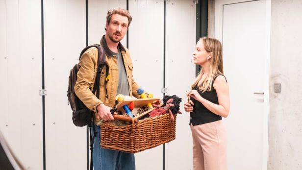 Ein Mann mit Rucksack hält einen Korb voller Spielzeug, neben ihm spricht eine Frau vor einer weißen Tür.
