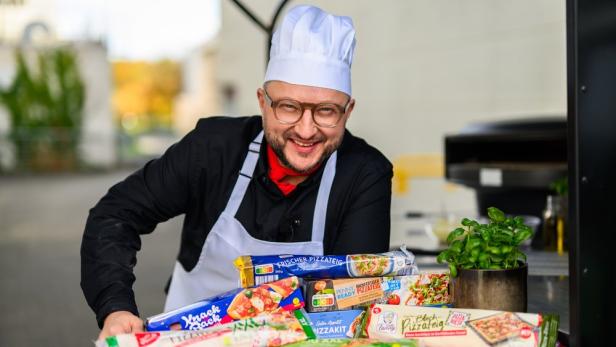 Ein lächelnder Koch mit weißer Mütze präsentiert verschiedene Packungen Pizzateig vor einem Pizzaofen.