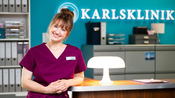 Lächelnde Mitarbeiterin in bordeauxfarbener OP-Kleidung an der Rezeption einer Klinik, mit türkisfarbener Wand und beleuchtetem Tresen.