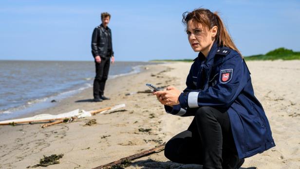 Polizistin kniet am Strand mit Handy, im Hintergrund steht ein Mann am Wasser.