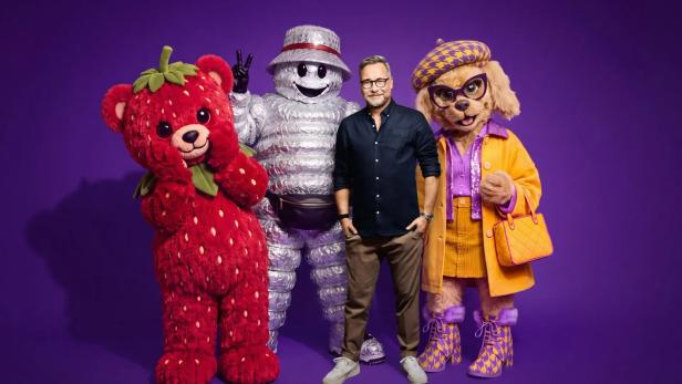 Matthias Opdenhövel steht vor einem violetten Hintergrund zwischen drei Maskottchen aus The Masked Singer.