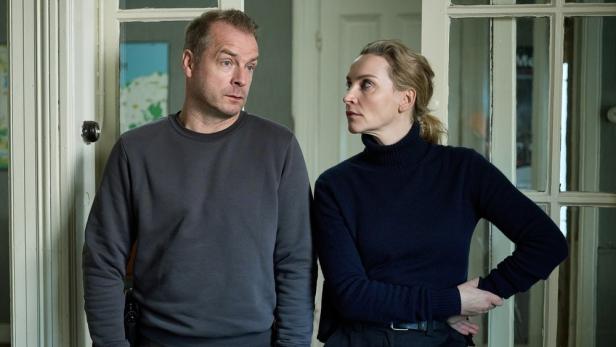 Ein Mann in einem grauen Pullover und eine Frau in einem dunkelblauen Rollkragenpullover stehen nebeneinander und schauen sich ernst an.
