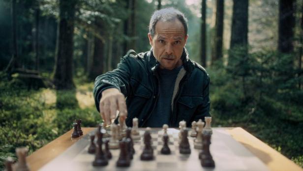 Ein Mann mit Schnurrbart spielt konzentriert Schach an einem Tisch inmitten eines Waldes.