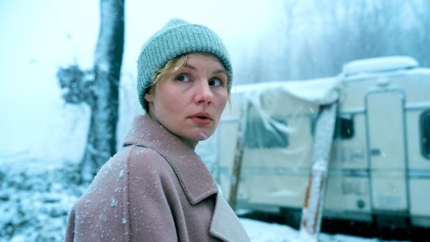 Eine Frau mit hellblauer Mütze und beigem Mantel blickt in einer verschneiten Winterlandschaft vor einem Wohnwagen besorgt zur Seite.