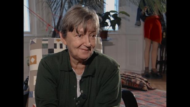 Christine Nöstlinger mit kurzem Haar und dunkelgrünem Pullover sitzt lächelnd in einem Sessel vor einem hellen Raum mit Pflanzen.