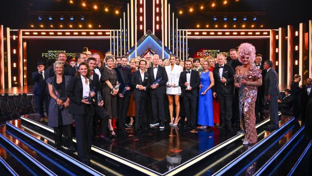 Preisträger posieren mit ihren Trophäen auf der Bühne bei der Verleihung des Deutschen Fernsehpreises vor einer beleuchteten Kulisse.