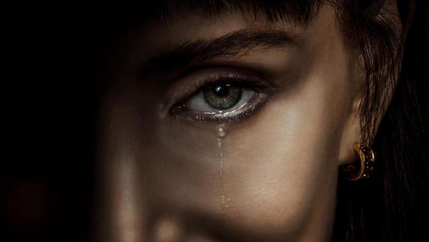 Nahaufnahme einer Frau mit einem rätselhaften Lächeln und einer Träne im Auge vor schwarzem Hintergrund mit dem Schriftzug „OTHER MOMMY“.