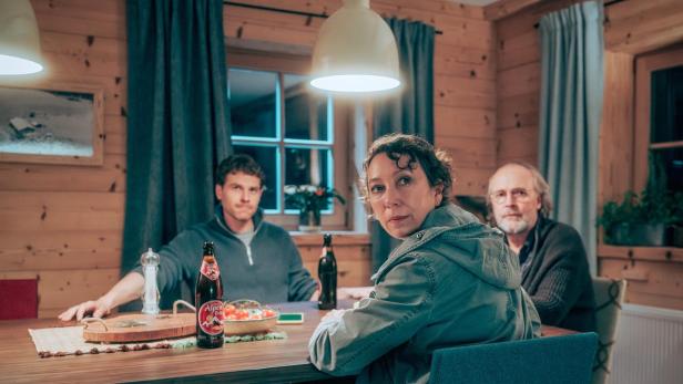 Drei Personen sitzen in einer gemütlichen Holzhütte an einem Tisch mit einer Flasche Alpen-Bier.