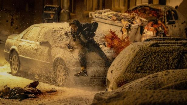 Mann springt in einer verschneiten Straßenszene vor Explosionen zwischen Autos.