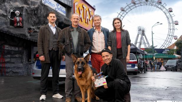 Das Team der Serie Kommissar Rex posiert mit einem Deutschen Schäferhund und einer Filmklappe neben einer Prater-Geisterbahn mit dem Wiener Riesenrad im Hintergrund.