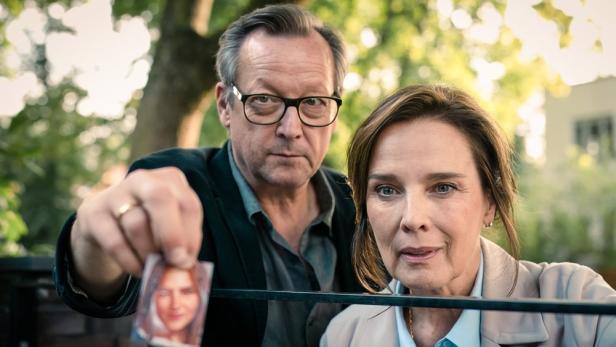 Ein Mann mit Brille und eine Frau schauen ernst in die Kamera, während er ein kleines Porträtfoto einer jungen Frau in die Höhe hält.
