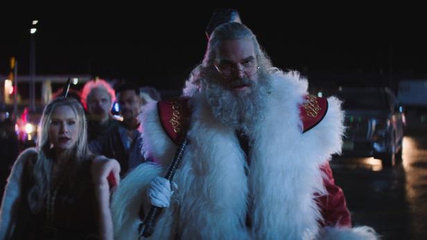 Ein als Weihnachtsmann gekleideter Mann mit Axt geht entschlossen durch die Nacht, gefolgt von einer Frau und weiteren Personen.