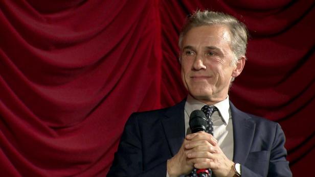Christoph Waltz spricht in einem Anzug mit Krawatte vor einem roten KIno-Vorhang in ein Mikrofon.