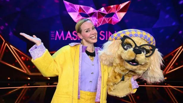 Eine lächelnde Frau posiert neben einer Hundekostüm-Maske auf einer bunten Masked-Singer-Bühne.