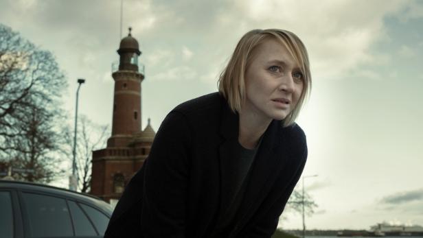 Eine blonde Frau blickt mit besorgtem Ausdruck vor einem Leuchtturm in die Ferne.
