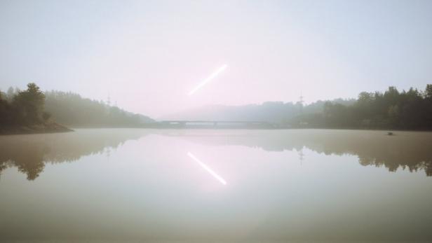Ein nebliger See mit einer Brücke im Hintergrund und einer hellen Lichtlinie am Himmel, die sich im ruhigen Wasser spiegelt.