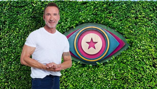 Jochen Bendel steht vor einer grünen Hecke mit dem ikonischen Big-Brother-Auge im Hintergrund.