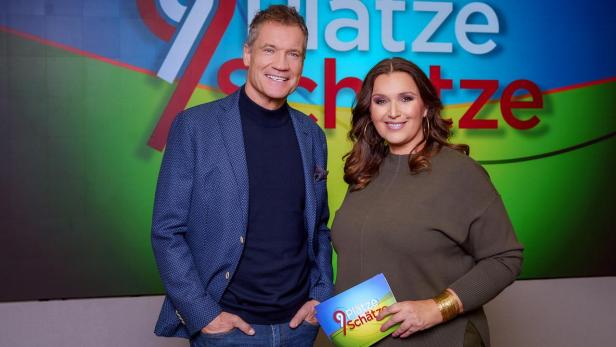 Die Moderatoren von „9 Plätze – 9 Schätze“ stehen lächelnd vor einem farbenfrohen Hintergrund mit dem Sendungslogo.