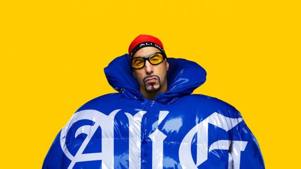 Ali G trägt eine überdimensionale blaue Daunenjacke mit der Aufschrift „Ali G Who Iz I?“ vor einem gelben Hintergrund.