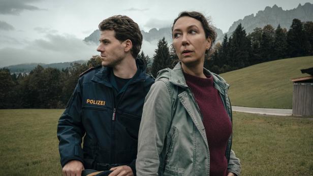 Polizist und Frau stehen auf einer Wiese vor Bergen und schauen in verschiedene Richtungen.