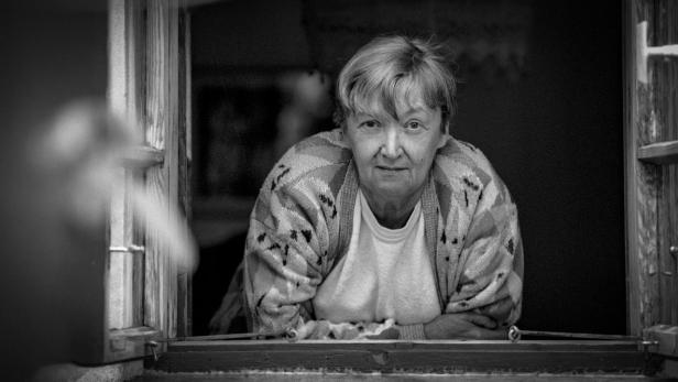 Eine ältere Frau mit kurzem Haar blickt in diesem schwarz-weiß Porträt direkt aus einem geöffneten Fenster nach draußen.