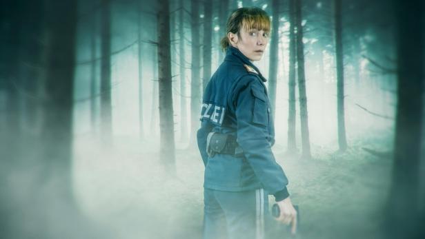 Eine Polizistin in Uniform steht mit einer Pistole in der Hand in einem nebligen Wald und blickt wachsam zurück.