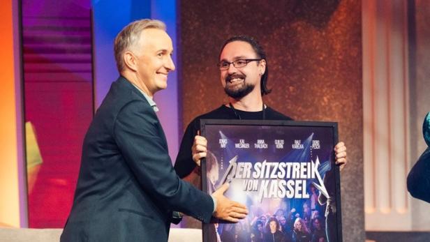 Zwei Männer lächeln, einer überreicht ein gerahmtes Plakat mit dem Titel „Der Sitzstreik von Kassel“.