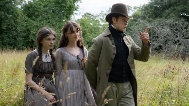 Ein Mann mit Zylinder und zwei Frauen in historischen Kleidern stehen in einer Wiese, während der Mann nach vorne zeigt.