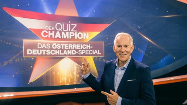 Johannes B. Kerner präsentiert vor einer Studiowand mit dem Logo von „Der Quiz-Champion – Das Österreich-Deutschland-Special“.