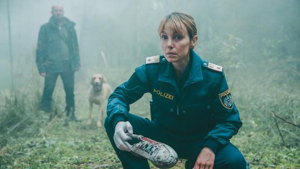 Eine Polizistin in dunkler Uniform hält im Wald einen blutigen Schuh, während im Hintergrund ein Mann mit einem Hund steht.
