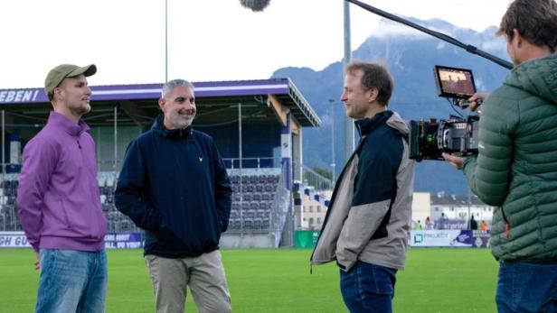 Ein Kamerateam filmt drei Männer im Gespräch auf einem Fußballplatz vor einer Tribüne.