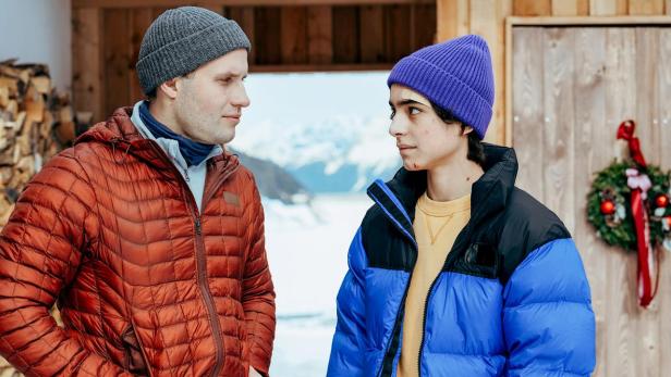 Ein Mann mit grauer Mütze und oranger Jacke blickt einen Jüngeren mit blauer Mütze und farbiger Jacke an, der leichte Verletzungen im Gesicht hat.