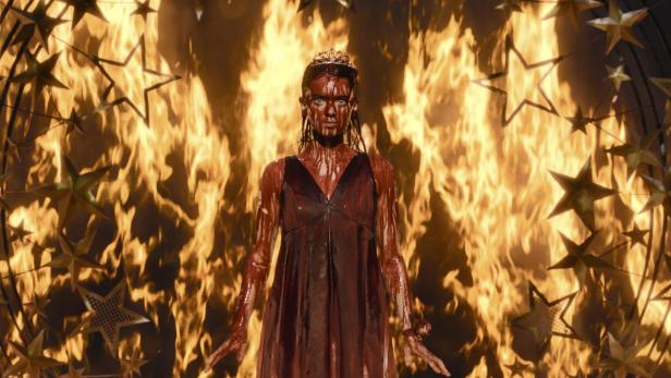 Eine blutverschmierte Frau im dunklen Kleid und mit Krone steht vor lodernden Flammen.