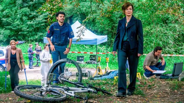 Einsatzkräfte und Polizisten untersuchen im Wald einen Tatort mit einem liegenden Fahrrad und abgesperrtem Bereich.