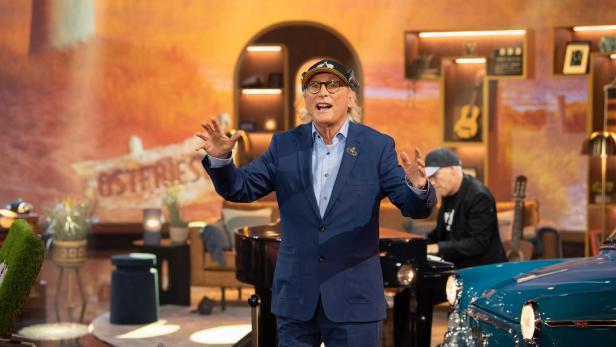 Otto Waalkes im blauen Anzug und mit Kappe spricht gestikulierend in einem TV-Studio, daneben sitzt ein Pianist am Flügel.