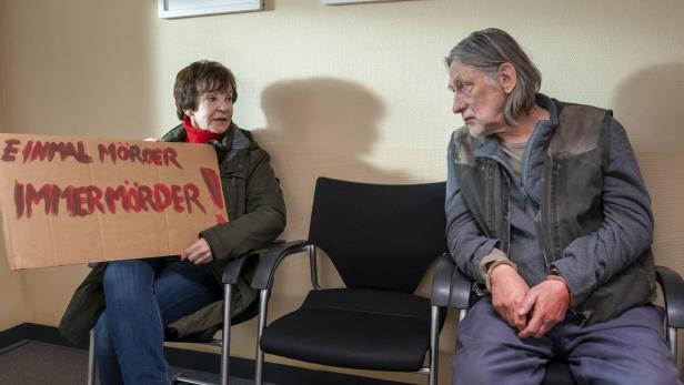 Zwei ältere Personen sitzen in einem Warteraum; die Frau hält ein Schild mit der Aufschrift „Einmal Mörder, immer Mörder“.