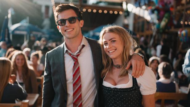 Ein lächelndes Paar steht Arm in Arm auf einem Fest; er trägt Anzug und Krawatte, sie ein traditionelles Dirndl.